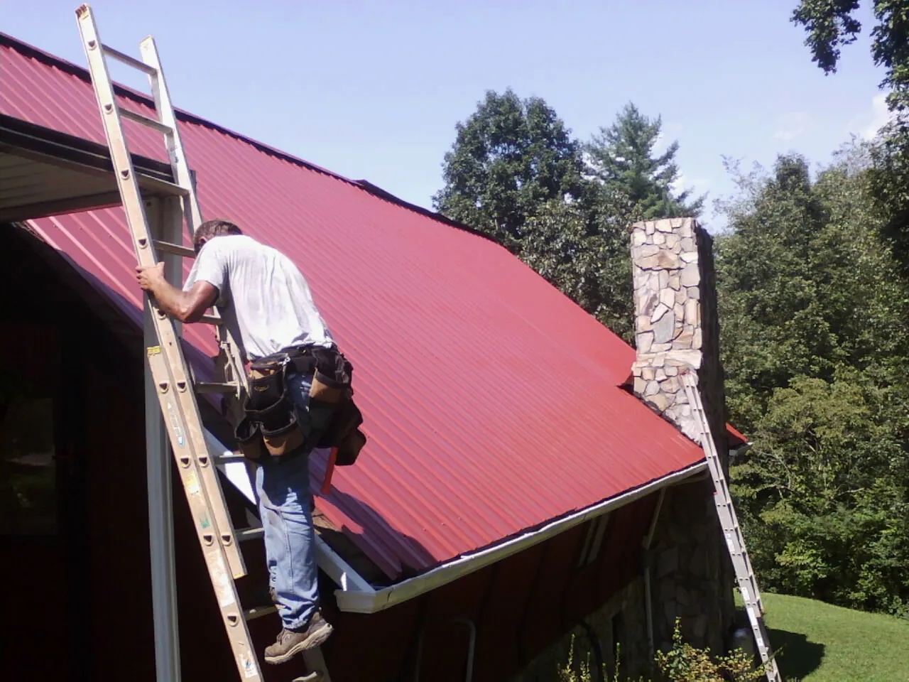 Roofer on a ladder beside a red metal roof and stone chimney — Soffit & Fascia Repair by Roofing Brothers in Harleyville